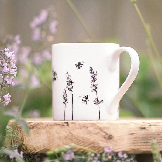 White mug with black floral design on a wooden surface surrounded by greenery and flowers