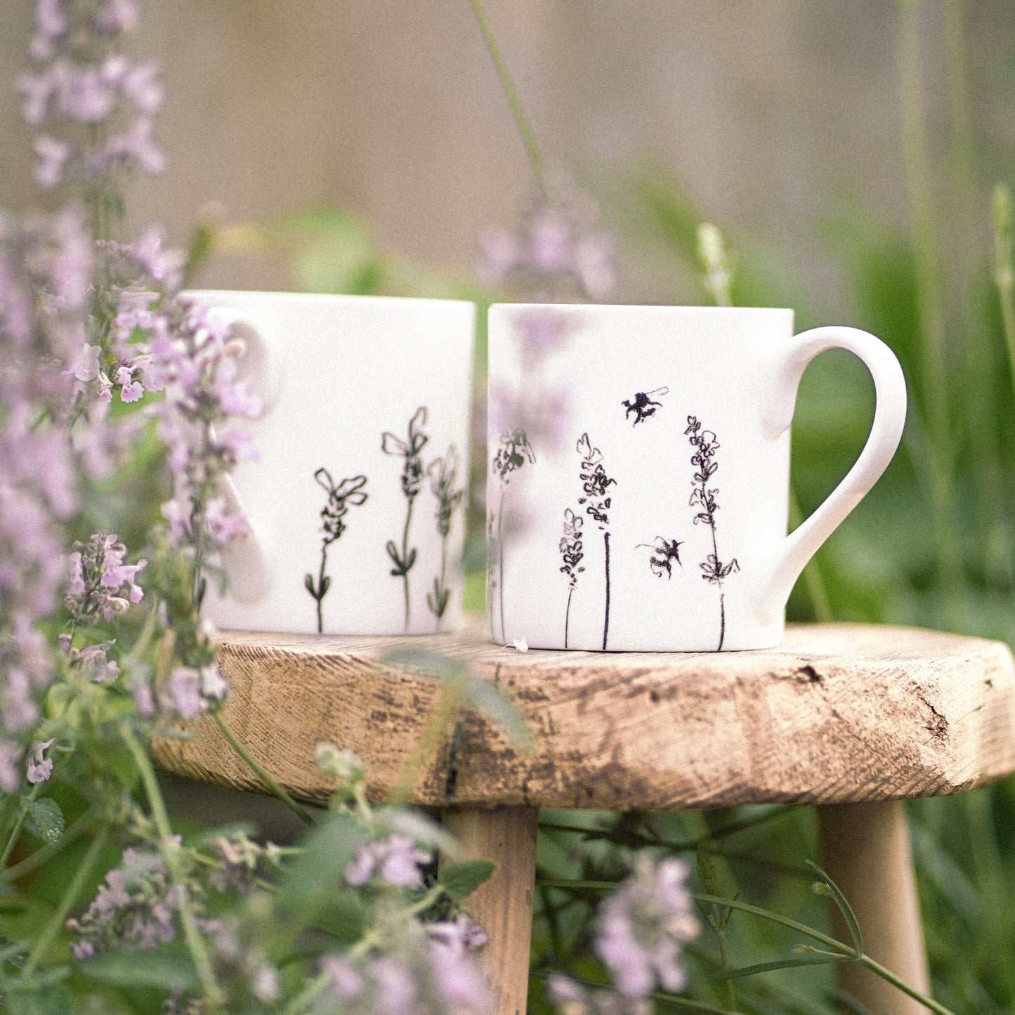 Two white mugs with floral designs on a wooden stool surrounded by flowers and greenery