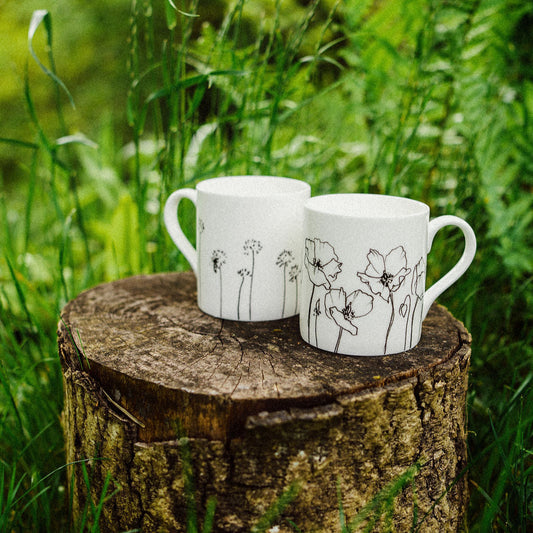 Two white mugs with floral designs on a wooden stump in a grassy outdoor setting