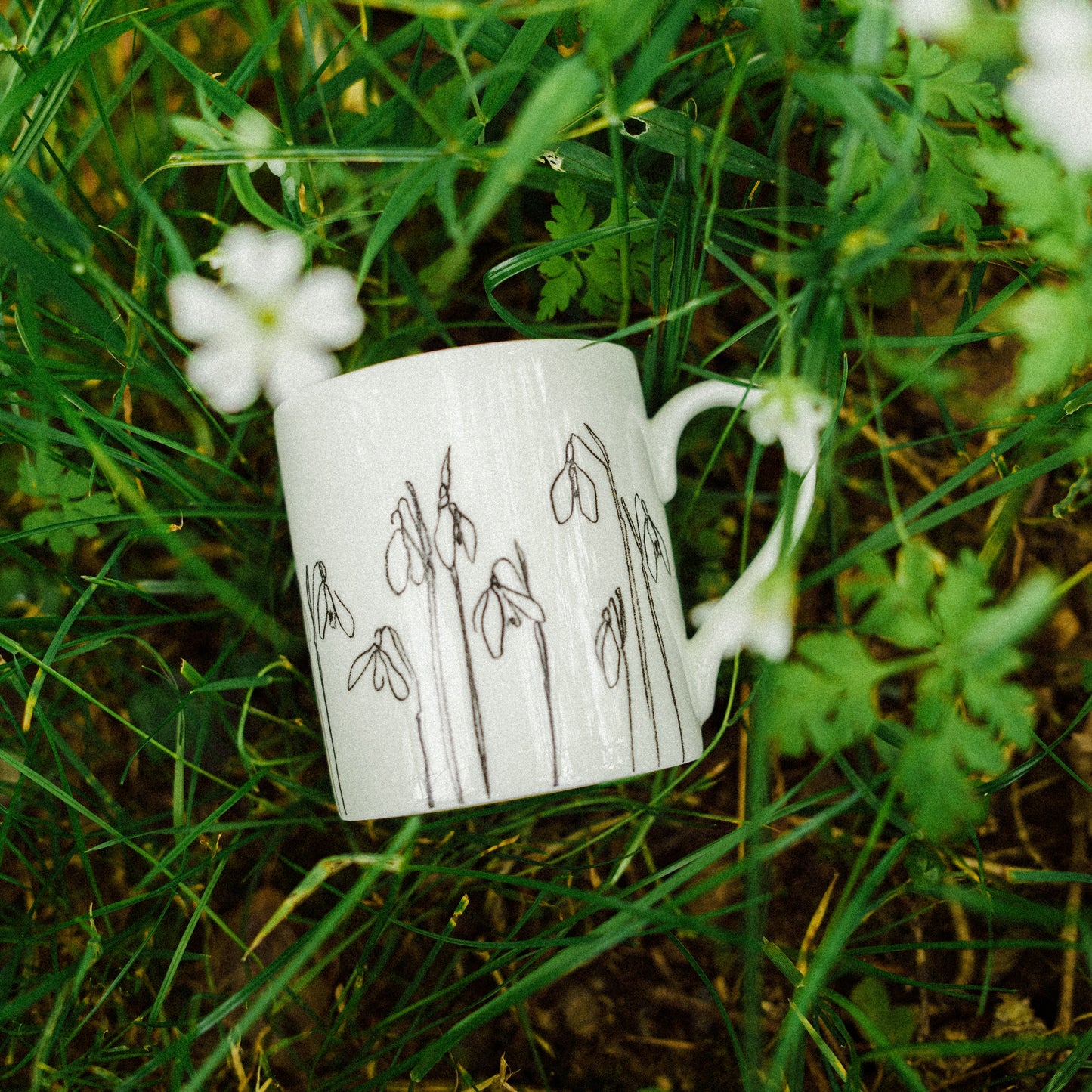 White mug with line drawings of flowers, laid on grassy background