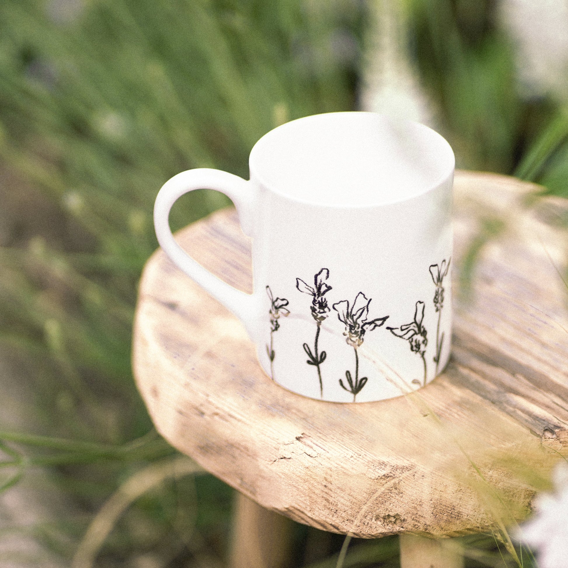 White mug with black floral design on a wooden stool surrounded by greenery