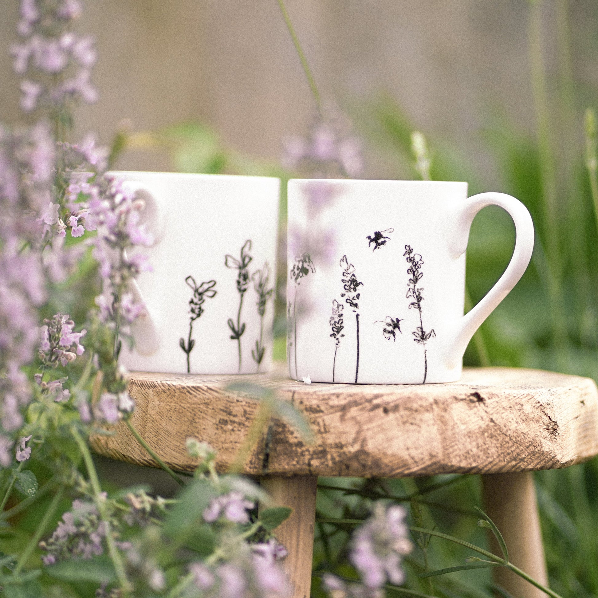Two white mugs with floral designs on a wooden stool surrounded by flowers and greenery