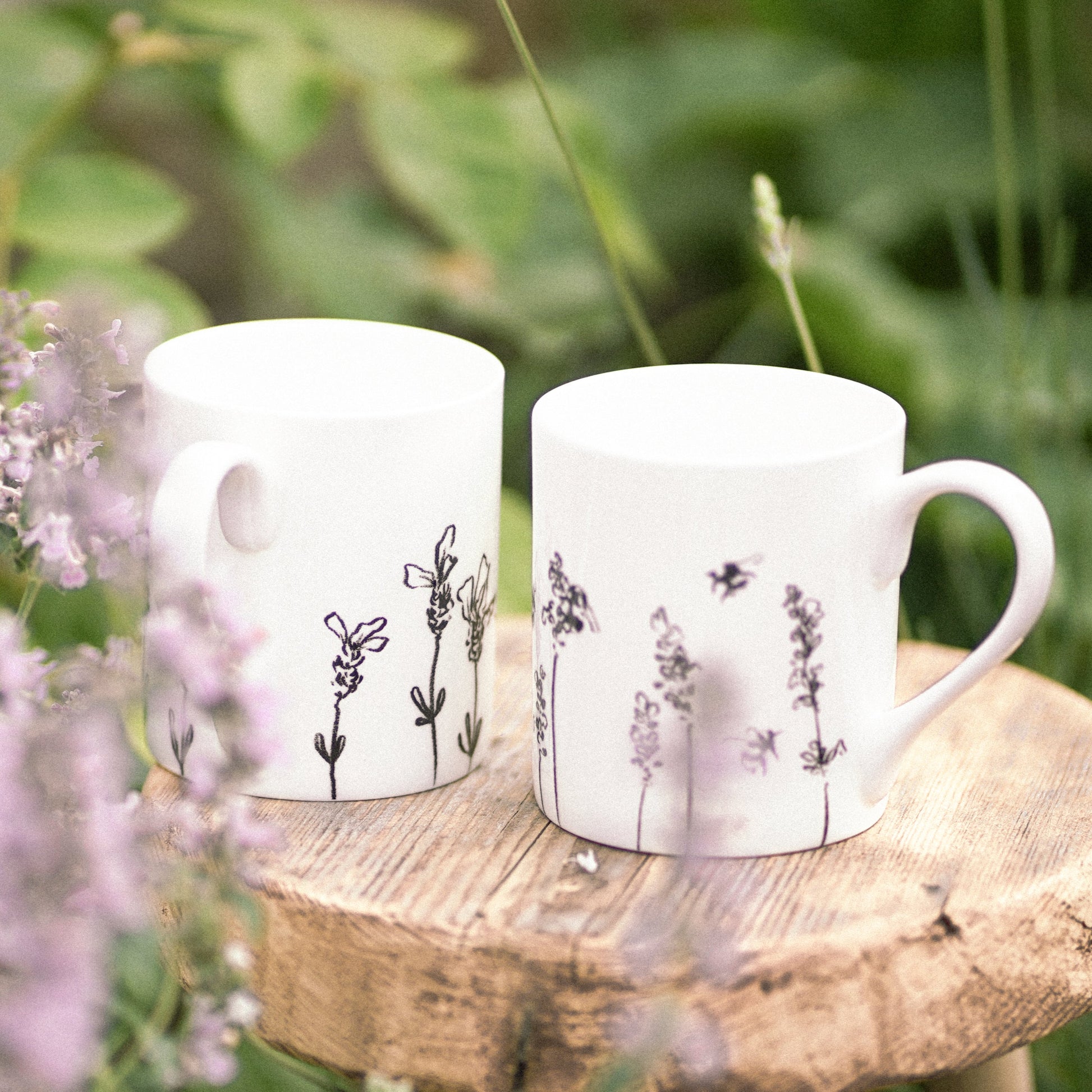 Two white mugs with floral designs on a wooden stool surrounded by flowers and greenery.