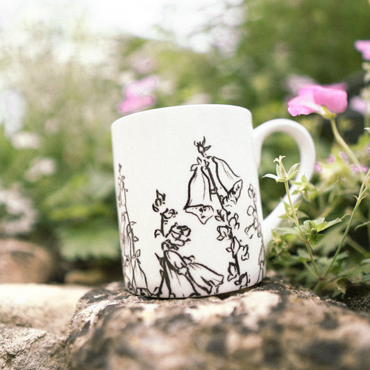 White mug with black illustrations on a stone surface with greenery and flowers in the background