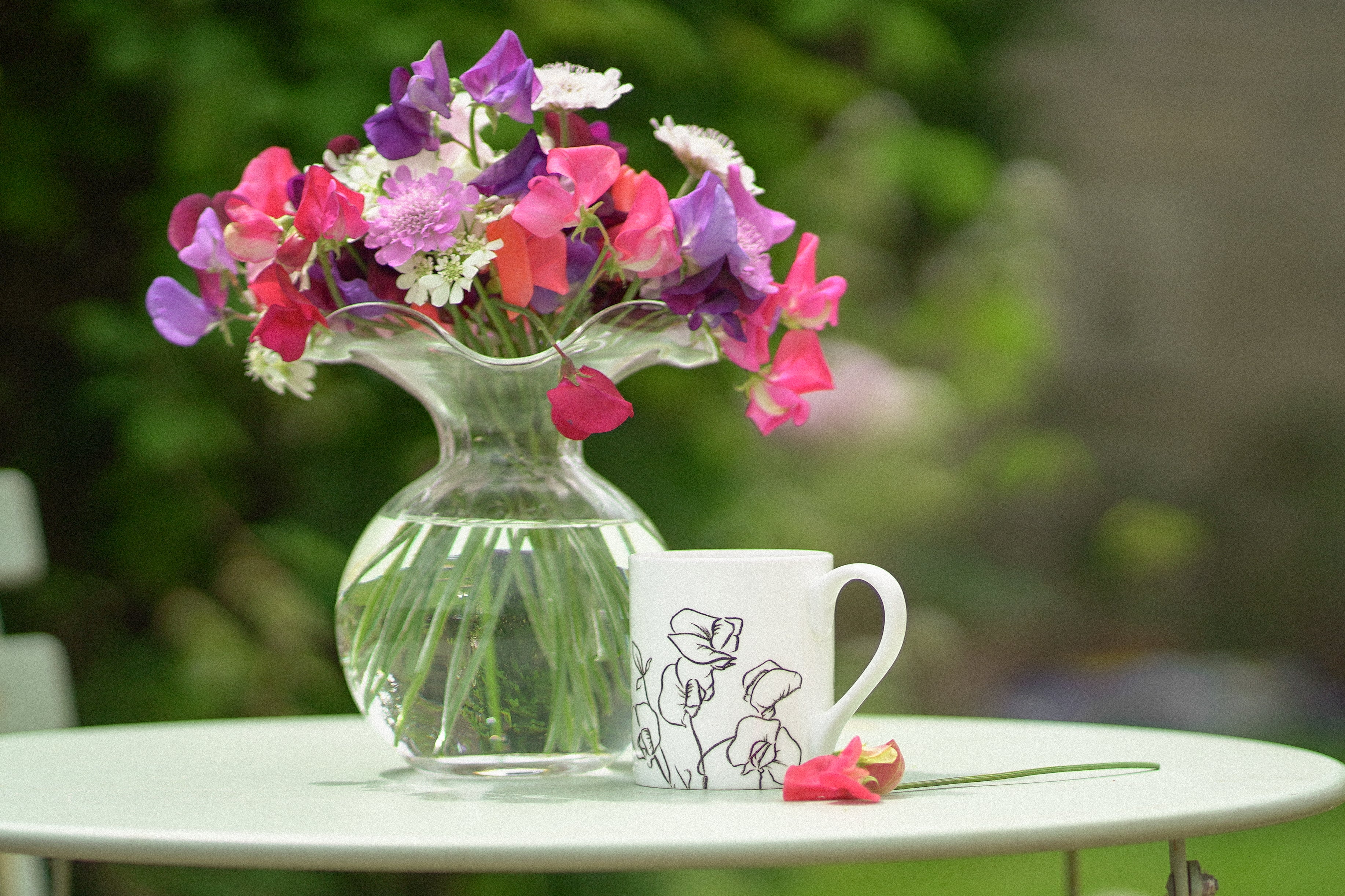 Clear vase with colorful flowers on a table next to a white mug with floral design.
