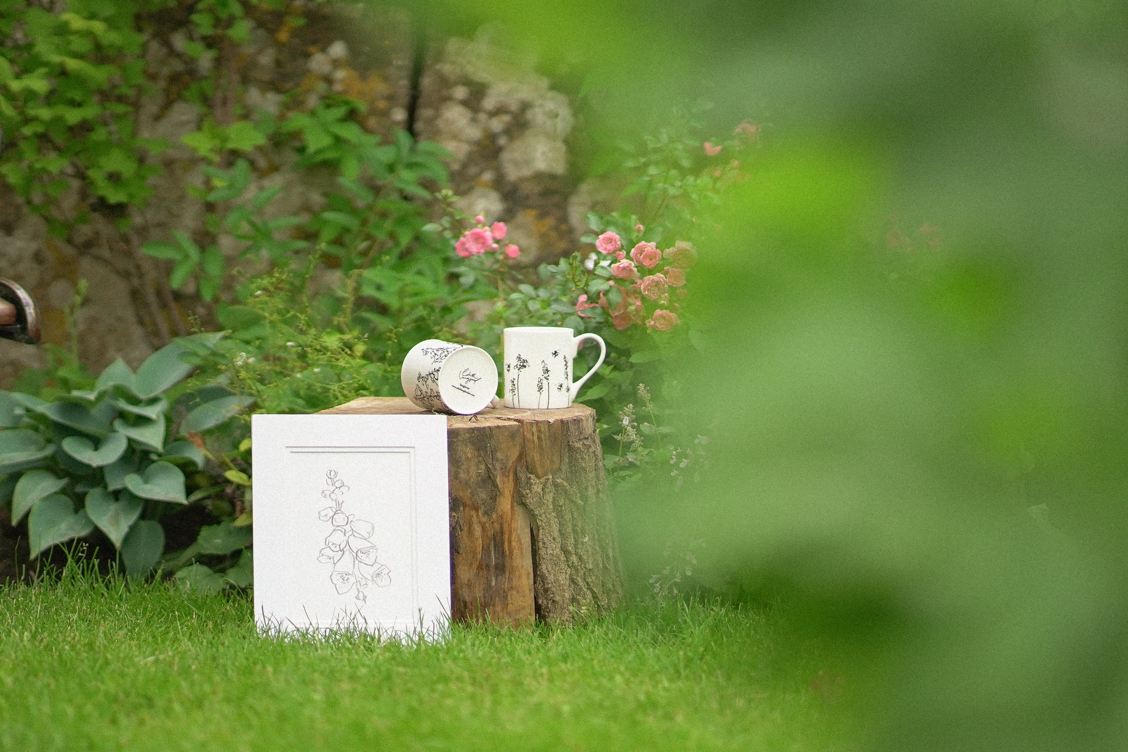 Two white mugs and a book on a wooden log in a garden setting with flowers and greenery.