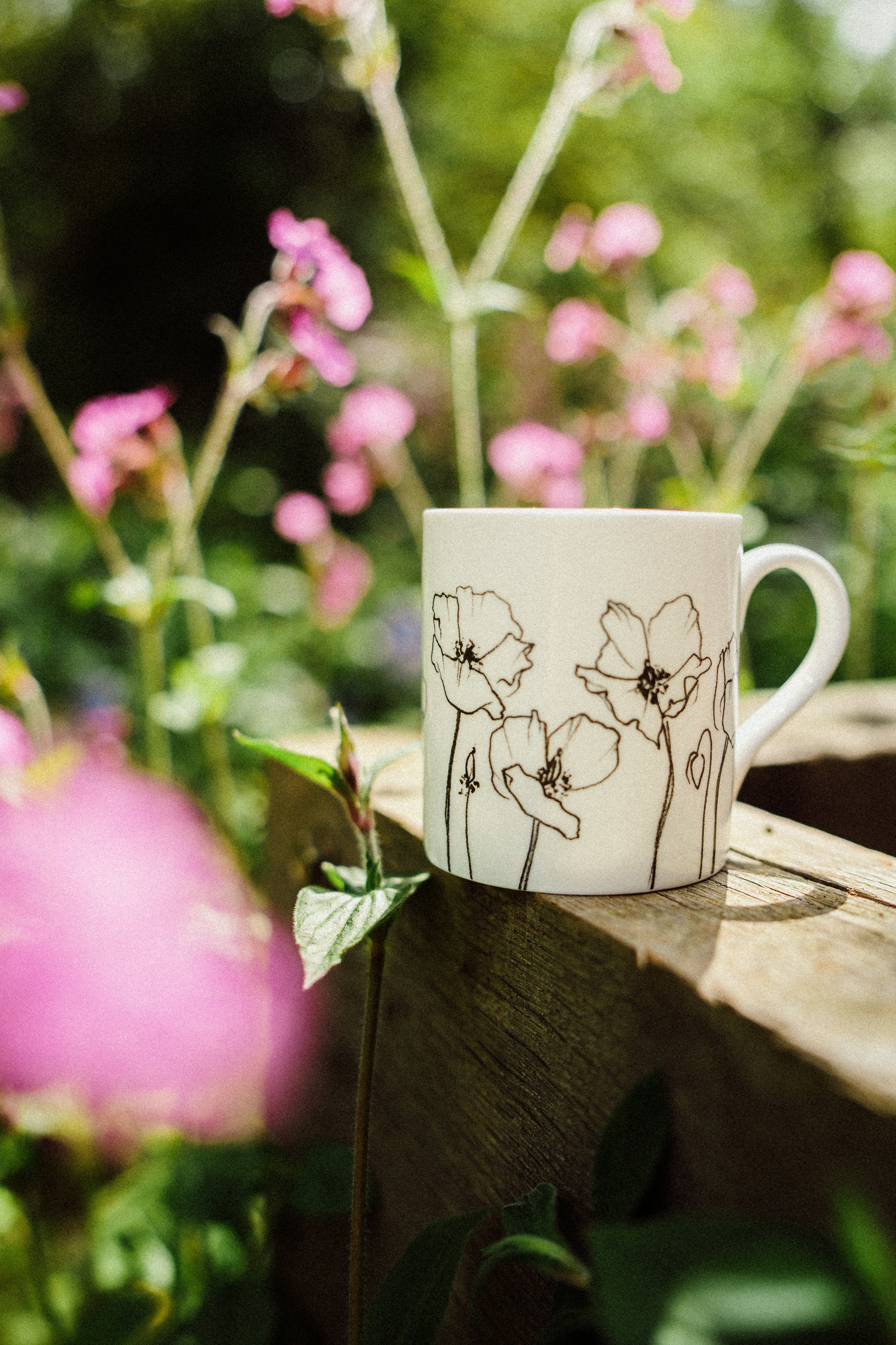 White mug with floral design on a wooden surface with pink flowers in the background