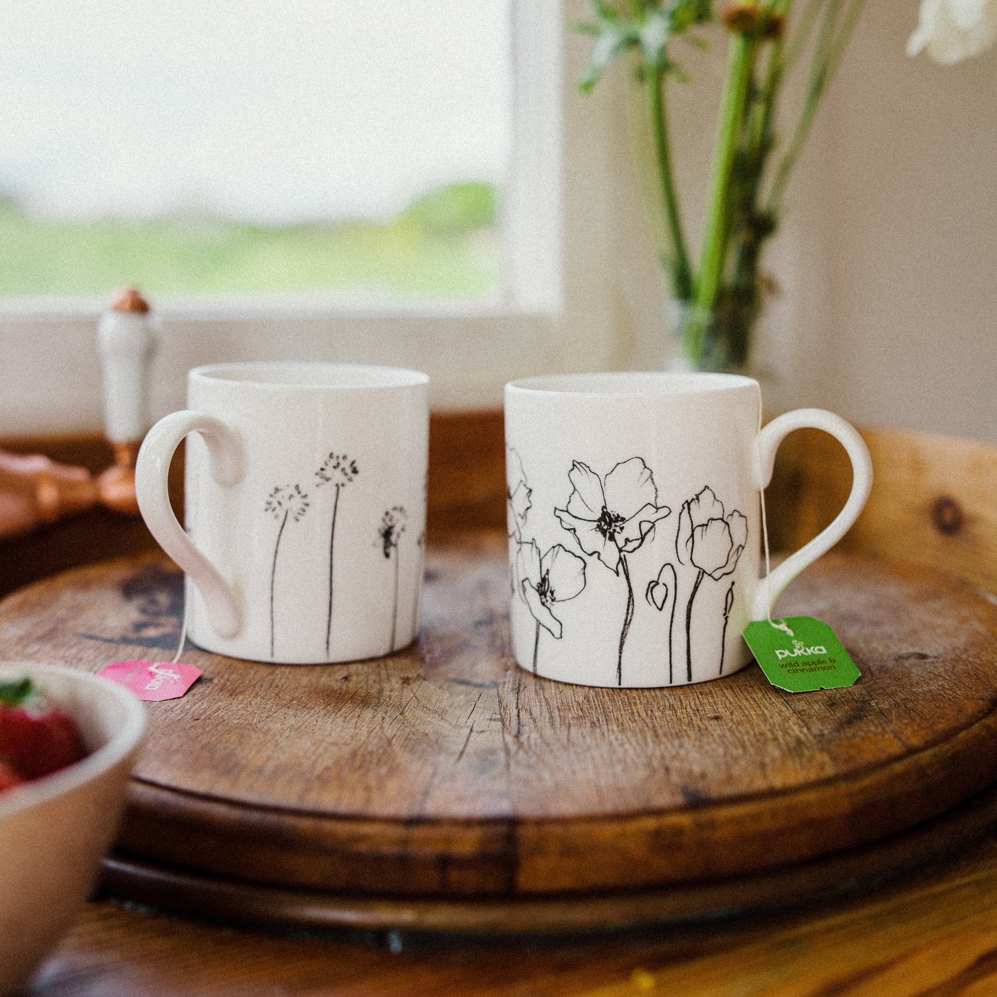 Two white mugs with black floral illustrations on a wooden surface