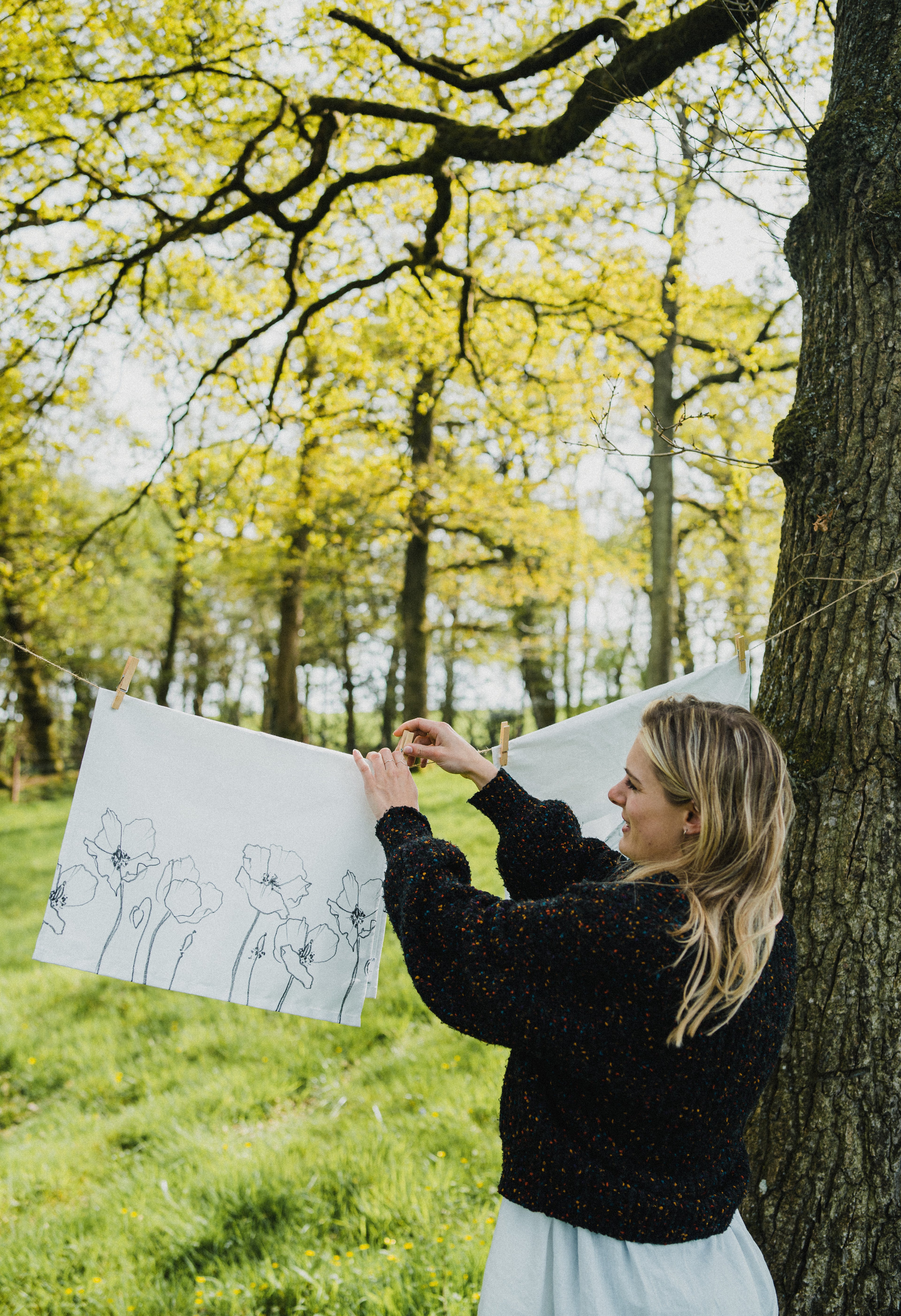 A woman in a black knit sweater and white skirt hangs a large white canvas print outdoors between trees in a grassy forest area. The canvas features black line-drawn floral illustrations.